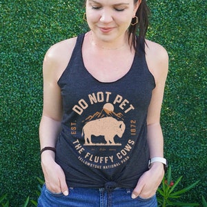 May include: A dark grey tank top with a white graphic print that says "Do Not Pet The Fluffy Cows Est. 1872 Yellowstone National Park". The graphic also features a brown silhouette of a bison in front of a mountain range.