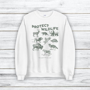 May include: White sweatshirt with a green graphic of various animals, including a deer, bear, and tiger. The text "PROTECT WILDLIFE" is printed above the animals, with "CULTURED COLLECTIVES" below. The sweatshirt has a crew neck and long sleeves.