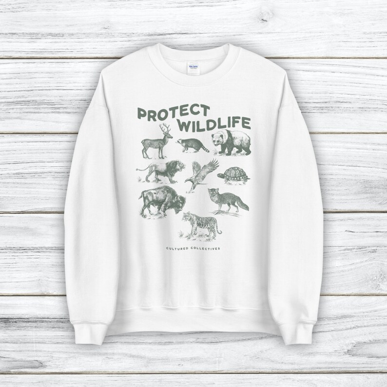 May include: White sweatshirt with a green graphic of various animals, including a deer, bear, and tiger. The text "PROTECT WILDLIFE" is printed above the animals, with "CULTURED COLLECTIVES" below. The sweatshirt has a crew neck and long sleeves.