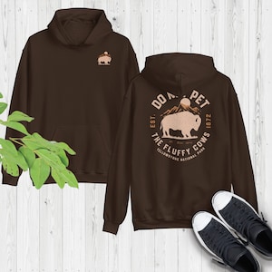 Może przedstawiać: Brązowa bluza z kapturem z nadrukiem na plecach, który głosi "Do Not Pet The Fluffy Cows, Est. 1872, Yellowstone National Park". Grafika przedstawia brązowego bizona przed pasmem górskim.
