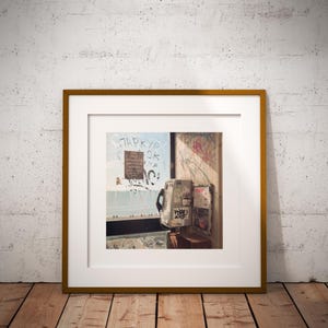 May include: Framed artwork featuring a vintage payphone with graffiti and a weathered appearance. The artwork is set against a light blue and white background with additional graffiti. The frame is a warm, golden brown.