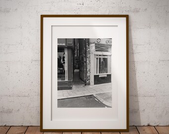 May include: Black and white photograph of a narrow street in a city. The street is lined with buildings, and there is a person walking down the street. The photo is framed in a brown frame.