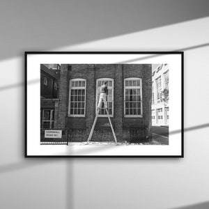 May include: Black and white framed photograph of a brick building with three large windows. A person is standing on a ladder, working on the center window. A street sign reads "Cornwall Road 361".