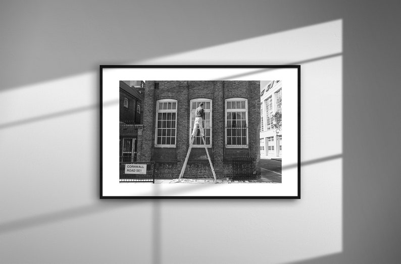 May include: Black and white framed photograph of a brick building with three large windows. A person is standing on a ladder, working on the center window. A street sign reads "Cornwall Road 361".