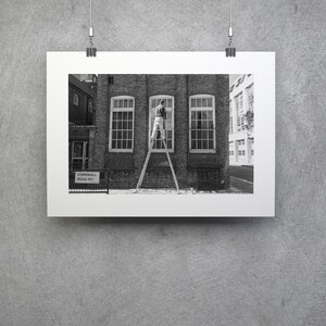 May include: Black and white photograph of a framed print. The print depicts a person on a ladder cleaning a window of a brick building. The building has three windows. A street sign is visible in the foreground.