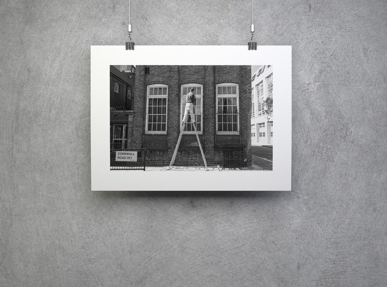 May include: Black and white photograph of a framed print. The print depicts a person on a ladder cleaning a window of a brick building. The building has three windows. A street sign is visible in the foreground.