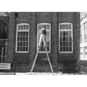 May include: Black and white image of a person on a ladder cleaning a window. The building is brick with three large windows. A street sign reads "Cornwall Road SE1".