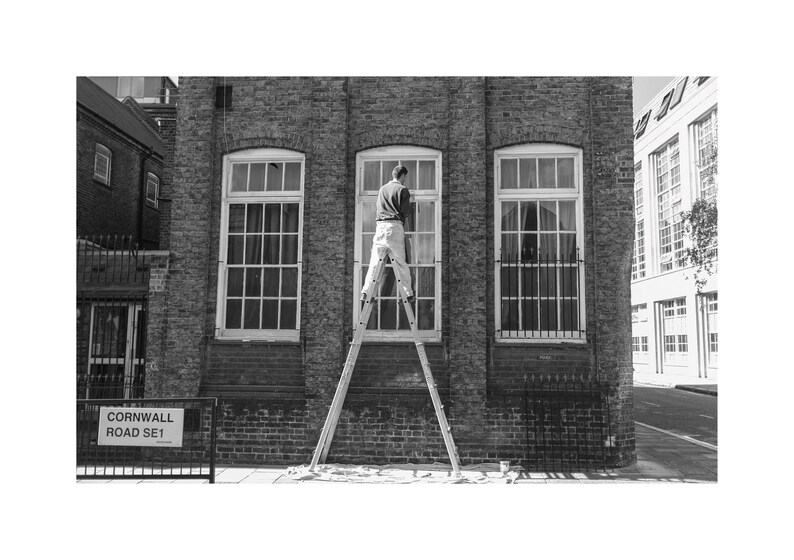 May include: Black and white image of a person on a ladder cleaning a window. The building is brick with three large windows. A street sign reads "Cornwall Road SE1".