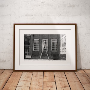 May include: A framed black and white photograph of a building with three large windows. A person is on a ladder, working on one of the windows. The frame is brown, and the photograph is displayed on a wooden floor against a white wall.