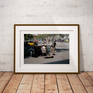 Può includere: Fotografia incorniciata di musicisti di strada che si esibiscono in una piazza della città. L'immagine mostra un pianista e un violinista che suonano in una giornata di sole, con pedoni e traffico sullo sfondo. La cornice è marrone e la fotografia è esposta su un pavimento di legno.