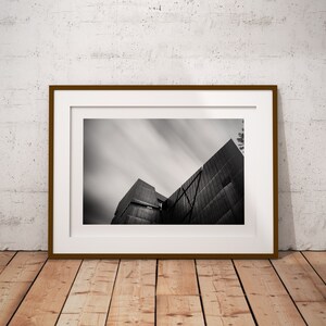 May include: Monochrome photograph of a contemporary building with angular architecture and a streaked sky, framed in a brown wooden frame with a white mount. The framed picture rests on a wooden floor.