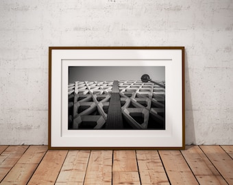 May include: Monochrome framed photograph of a building's geometric facade. The image is in a brown wooden frame, set against a white wall and wooden floor.