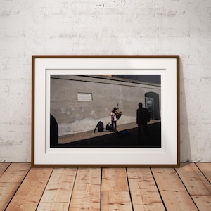 Può includere: Fotografia incorniciata di una scena di strada con un violinista che suona. L'immagine è ambientata in uno stretto vicolo con un muro strutturato. Una persona è in primo piano e un'altra sullo sfondo. La cornice è marrone.