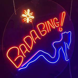 Può includere: Un cartello al neon con le parole "Badabing!" in rosso e arancione, con un contorno blu al neon delle gambe di una donna e una stella gialla sopra il testo.