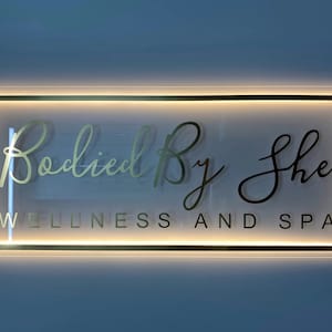 Puede incluir: Un letrero iluminado con marco negro y fondo blanco. El letrero dice "Bodied By She Wellness and Spa" en una fuente cursiva dorada.