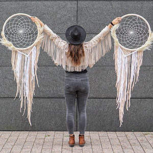 May include: A person wearing a black hat and a fringed jacket holds two large white dreamcatchers with feathers and tassels.