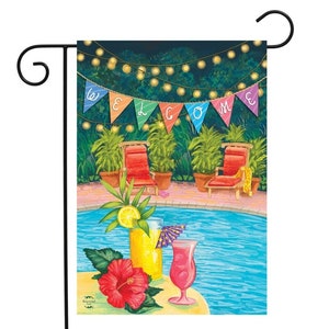 May include: A colorful welcome banner hangs above a swimming pool with two lounge chairs and string lights.  The pool is surrounded by tropical plants.  Two cocktails sit on a table with a red hibiscus flower.