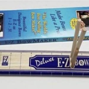 May include: A blue and white tool holder with the text "Deluxe E-Z Bow Maker" and a ruler with markings. The tool holder is made of wood and has a blue and white paint finish. The ruler is made of wood and has a white paint finish. Two wooden dowels are inserted into the tool holder.