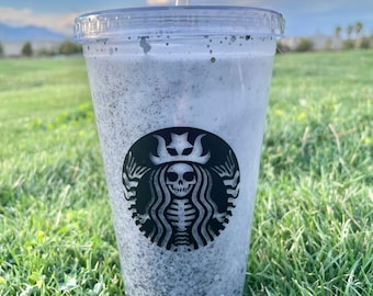May include: A clear plastic cup with a black and white Starbucks logo featuring a skeleton. The cup is filled with a white liquid and has a black straw.