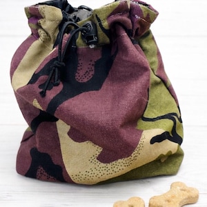 May include: A camouflage-patterned drawstring pouch in shades of green, burgundy, black, and beige. Two bone-shaped dog biscuits are in front of the pouch. The pouch has a black drawstring closure.