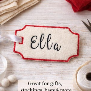 Op de afbeelding: Crèmekleurig vilten naamlabel met een rode rand en de naam "Ella" in zwart script. Een wit lint is bevestigd. De tekst onderaan luidt "Great for gifts, stockings, bags & more."