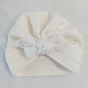 May include: White turban headband with a knotted bow on top. The headband is made of a soft, textured fabric.