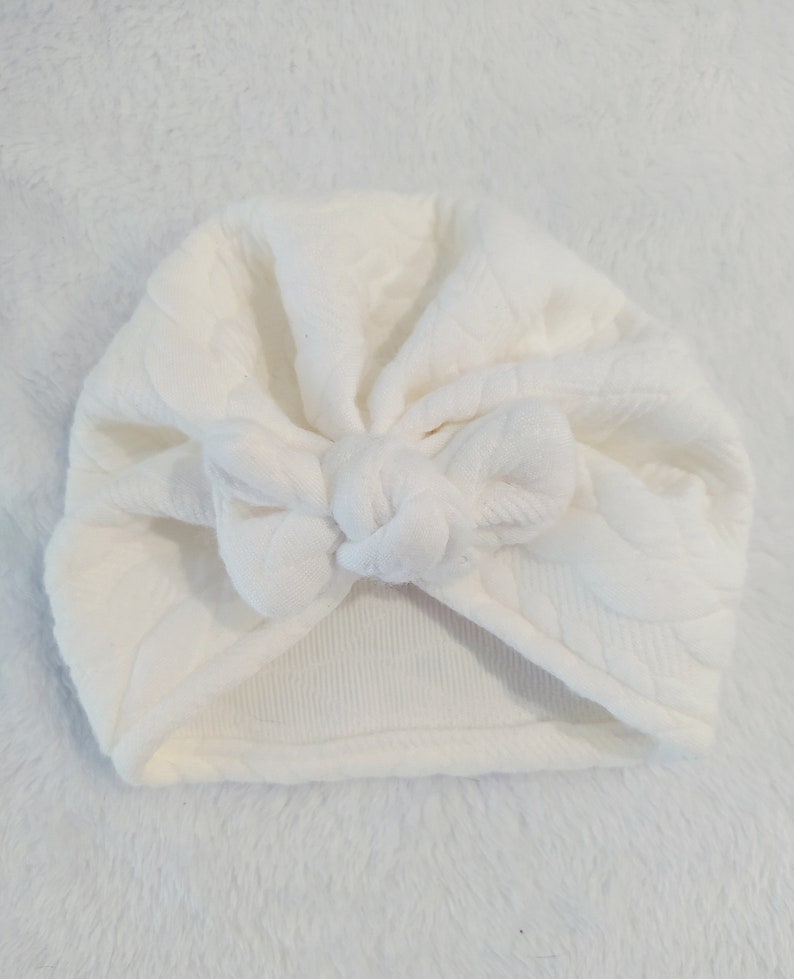 May include: White turban headband with a knotted bow on top. The headband is made of a soft, textured fabric.