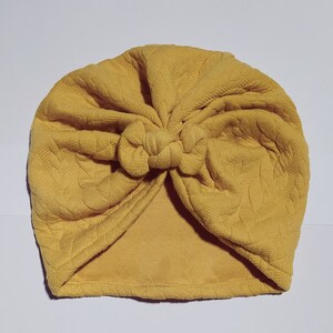 May include: A yellow turban with a knotted top. The turban is made of a soft, textured fabric.