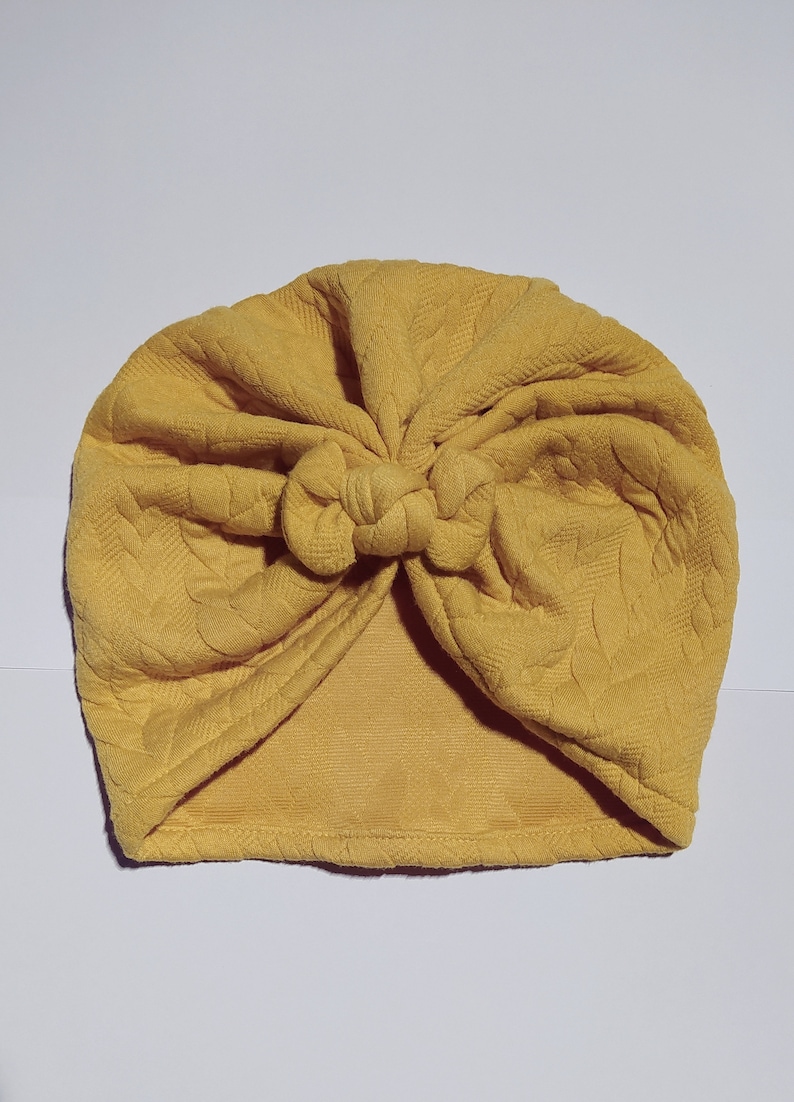 May include: A yellow turban with a knotted top. The turban is made of a soft, textured fabric.