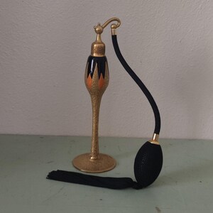 May include: An antique perfume atomizer with a gold-colored base and accents. The atomizer has a black and orange design on the bottle and a black bulb with a tassel. The atomizer is a vintage item.