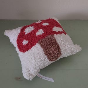 K&ouml;nnte beinhalten: Ein dekoratives Kissen mit einem Pilzdesign. Der Pilzhut ist rot mit wei&szlig;en Punkten und der Stiel ist braun. Das Kissen hat einen wei&szlig;en, strukturierten Hintergrund.
