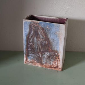 May include: Rectangular ceramic vase with a relief design. The vase features a blue and brown color scheme with a depiction of a figure and a crescent moon. The interior is a deep purple color. The vase is displayed on a green surface.