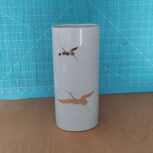 Puede incluir: Jarrón de cerámica blanca con borde dorado y dos grullas doradas en vuelo. El jarrón está sobre una superficie de madera.