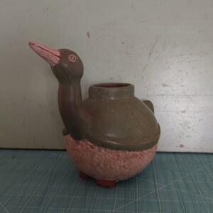 Pode incluir: Um vaso de cerâmica verde e marrom em forma de pássaro, com corpo redondo e pescoço longo. O vaso tem uma pequena abertura no topo para plantar.