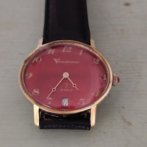 Könnte beinhalten: Eine goldfarbene Armbanduhr mit rotem Zifferblatt und schwarzem Lederarmband. Das Zifferblatt hat goldfarbene Stundenmarkierungen und Zeiger, und der Text "Vendome" und "7 Jewels" ist auf dem Zifferblatt aufgedruckt. Das Datumsfenster zeigt die Zahl "22".