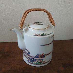 Puede incluir: Tetera de porcelana blanca con asa de mimbre marrón. La tetera tiene un diseño floral azul y blanco con un pájaro rojo.