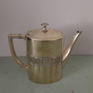 May include: A vintage silver-toned teapot with a geometric design band around the middle. The teapot has a curved handle, a lid with a small knob, and a long, slender spout. The teapot is likely made of metal.