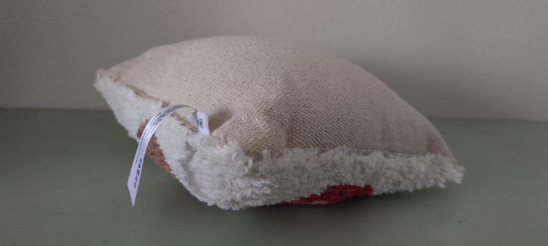 K&ouml;nnte beinhalten: Ein kleines, quadratisches Kissen mit einer strukturierten, cremefarbenen Stoffoberfl&auml;che und einem flauschigen, wei&szlig;en Rand. Die Unterseite des Kissens hat ein rotes Muster. Ein wei&szlig;es Etikett ist an der Seite befestigt.