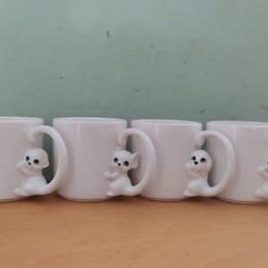 May include: Set of four white ceramic mugs with cat-shaped handles. Each mug features a white cat with black eyes and a pink nose.