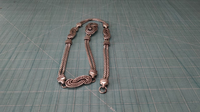 Peut inclure: Un collier en cha&icirc;ne argent&eacute;e avec un design d&eacute;coratif. La cha&icirc;ne a plusieurs brins et pr&eacute;sente des breloques orn&eacute;es en forme de paisley.