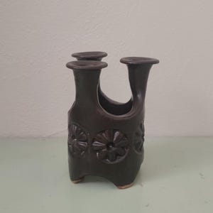 May include: A dark brown ceramic vase with a unique multi-spout top and three distinct openings. The cylindrical body features a repeating embossed floral pattern around the center and stands on three small, integrated feet at the base.
