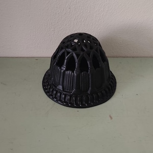 May include: A black, decorative flower frog with a domed shape and a series of arched openings. The base has a detailed, textured design. This item is likely used for arranging flowers.