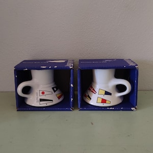 May include: Two white ceramic candle holders with blue and red nautical flag designs. The candle holders are in their original blue boxes.