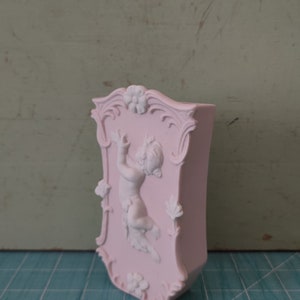 Puede incluir: Un jarr&oacute;n de cer&aacute;mica rosa con un dise&ntilde;o en relieve de un querub&iacute;n sosteniendo una flor. El jarr&oacute;n tiene un marco decorativo y ornamentado alrededor del querub&iacute;n.