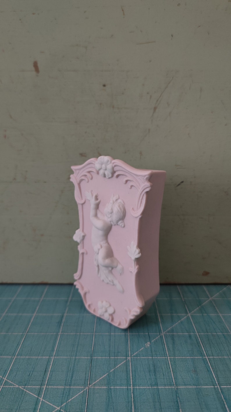 Puede incluir: Un jarr&oacute;n de cer&aacute;mica rosa con un dise&ntilde;o en relieve de un querub&iacute;n sosteniendo una flor. El jarr&oacute;n tiene un marco decorativo y ornamentado alrededor del querub&iacute;n.