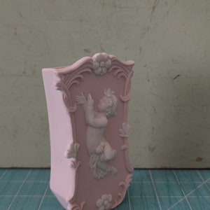 Puede incluir: Un jarr&oacute;n de cer&aacute;mica rosa con un dise&ntilde;o ornamentado que presenta una figura de querub&iacute;n blanco en relieve. El jarr&oacute;n tiene una parte superior festoneada y un motivo floral.