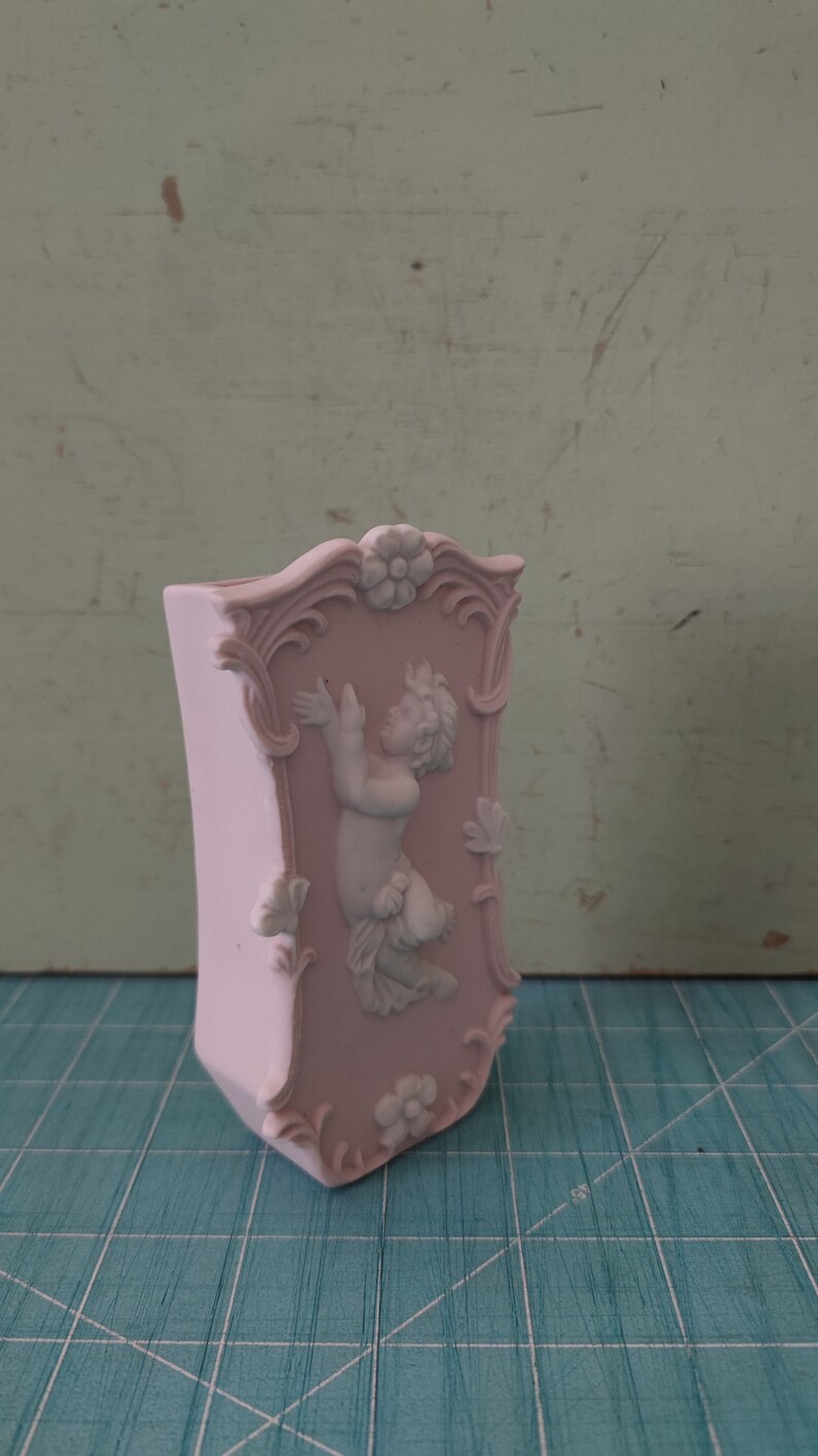 Puede incluir: Un jarr&oacute;n de cer&aacute;mica rosa con un dise&ntilde;o ornamentado que presenta una figura de querub&iacute;n blanco en relieve. El jarr&oacute;n tiene una parte superior festoneada y un motivo floral.