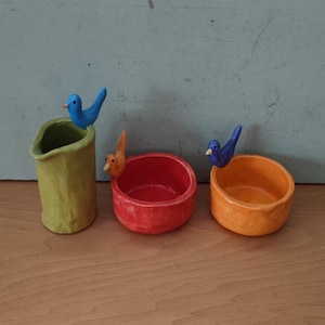 May include: Three colorful ceramic bird-topped containers. A green cylindrical container, a red bowl, and an orange bowl. Each has a small bird figurine perched on the edge. The background is a light blue wooden surface.