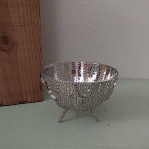 May include: A small, silver-colored metal bowl with a textured, perforated surface pattern. The bowl features a rounded shape and is supported by three thin, wavy metal legs that elevate it slightly above the surface it rests upon.