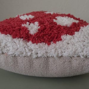 K&ouml;nnte beinhalten: Ein dekoratives kissenf&ouml;rmiges Kissen in Pilzform mit einem rot-wei&szlig;en getufteten Design. Die Oberseite des Kissens hat ein rotes Muster mit wei&szlig;en Akzenten, w&auml;hrend die R&auml;nder wei&szlig; sind. Das Kissen steht auf einer neutralfarbenen Stoffbasis.